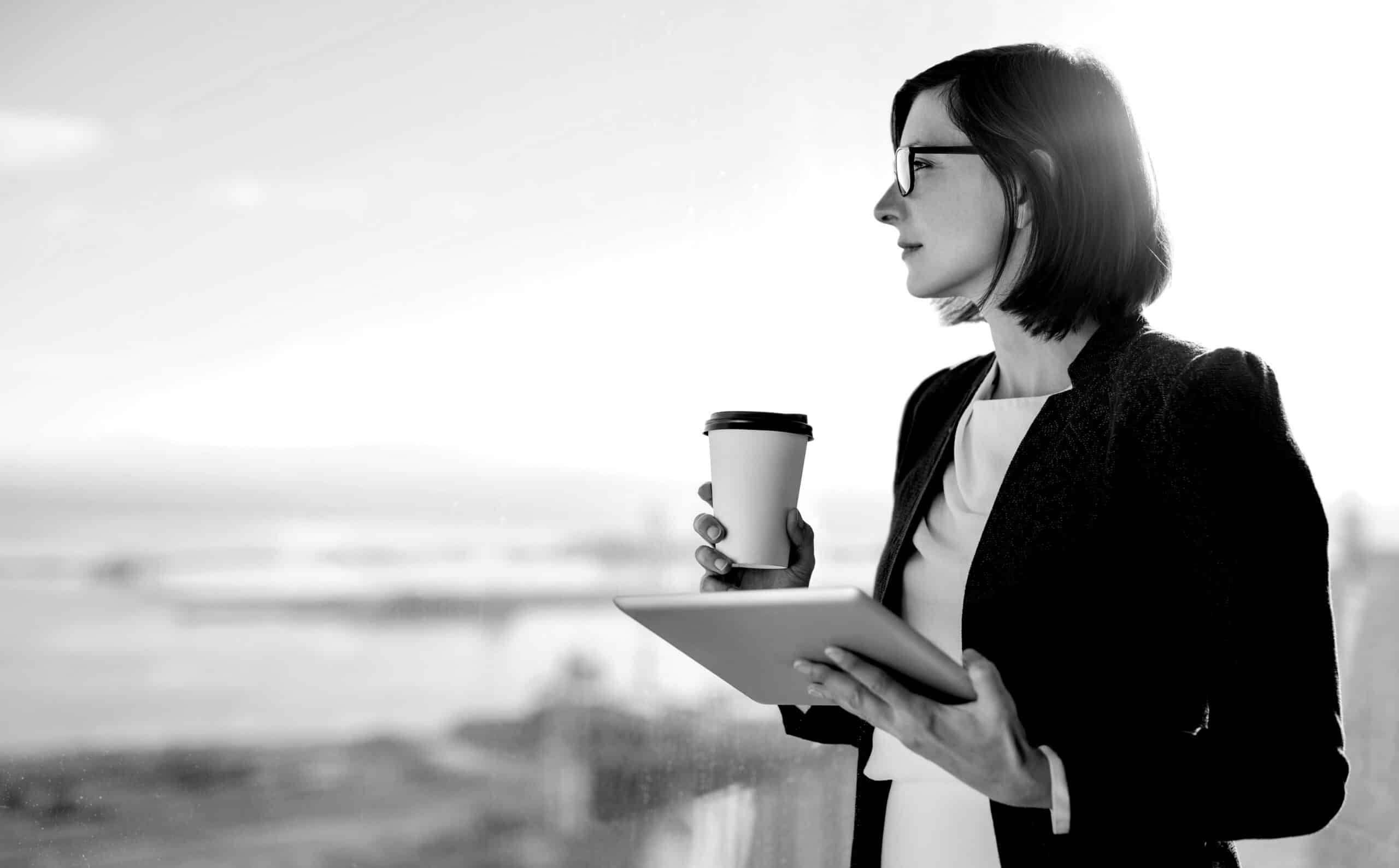 Attractive businesswoman drinking a coffee and using a digital tablet while standing at a window in an office overlooking the city